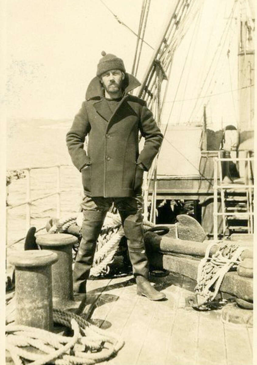 A man in heavy coat, tall boots, and a warm hat stands on the deck of a wooden ship, surrounded by ropes and nautical equipment, with the sea visible in the background.