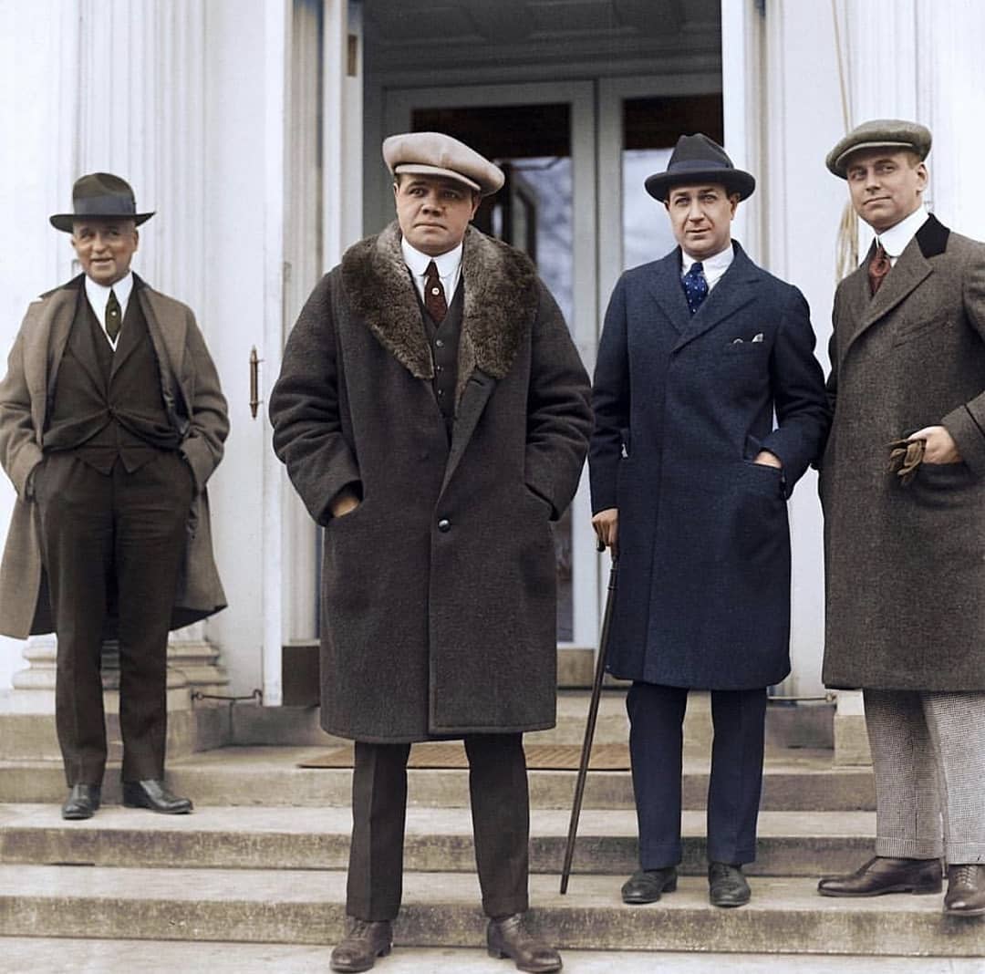 Four men in early 20th-century attire stand on steps in front of a building. They wear overcoats, hats, and dress shoes; one holds a cane. The mood appears formal and the setting is likely official or important.