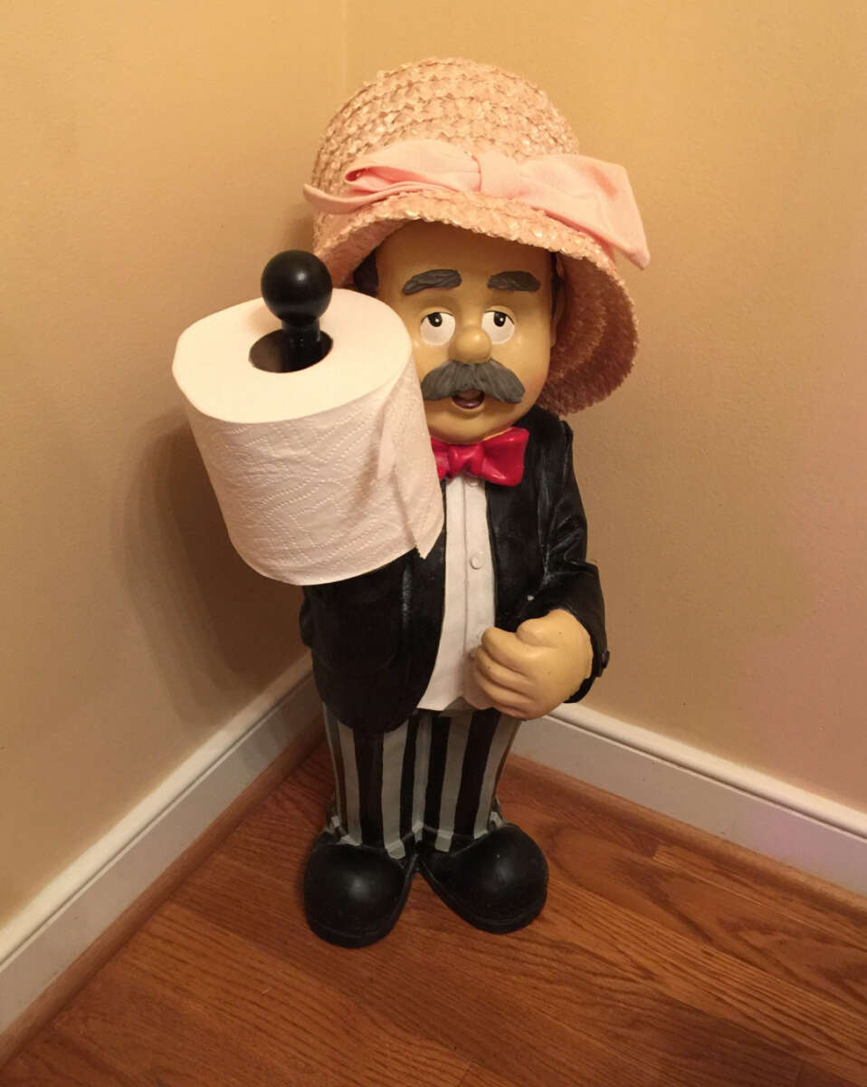 A whimsical statue of a man in striped pants, a black jacket, red bow tie, and a pink hat holding a roll of toilet paper on his outstretched arm, placed in the corner of a room with wooden flooring.