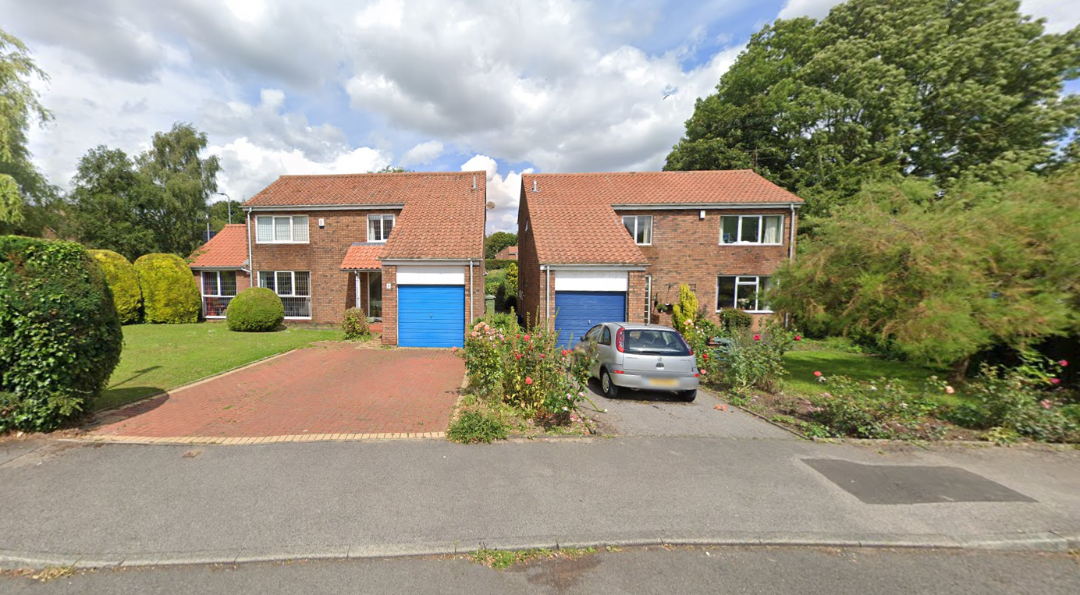 Two brick houses with red-tiled roofs and blue garage doors. A silver car is parked in front of the right house. Green lawns, bushes, and trees surround the houses under a partly cloudy sky.
