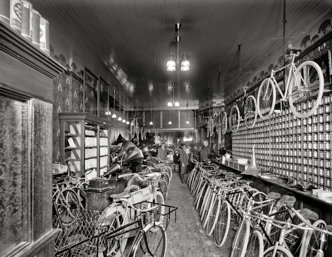 Black-and-white image of a vintage bicycle shop filled with bicycles, some hanging from the ceiling, and shelves lined with parts. Several people are standing and working at the back of the store.