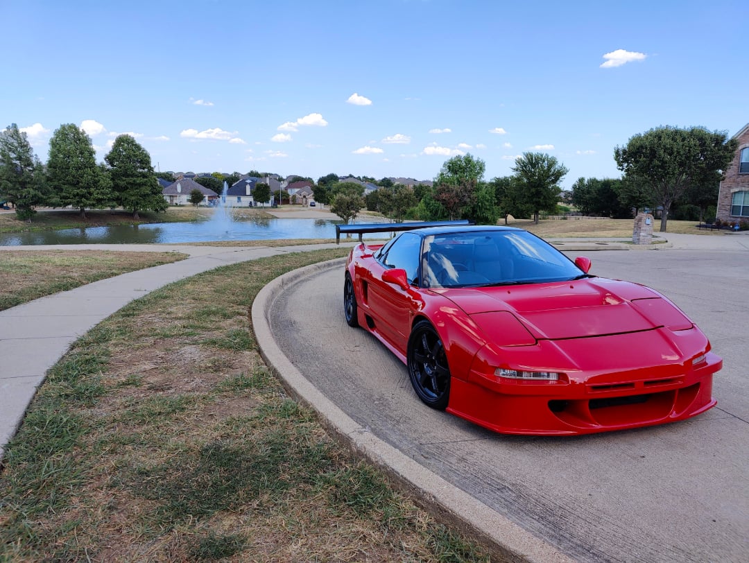 A red sports car is parked on a suburban street near a pond, with houses, trees, and a blue sky with scattered clouds in the background.