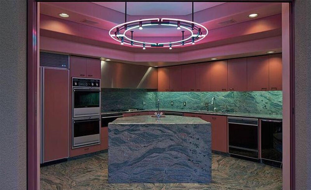 Modern kitchen with pink cabinets and a green marble island and backsplash. A large circular neon light fixture with pink and purple hues hangs from the ceiling, creating a retro ambiance.