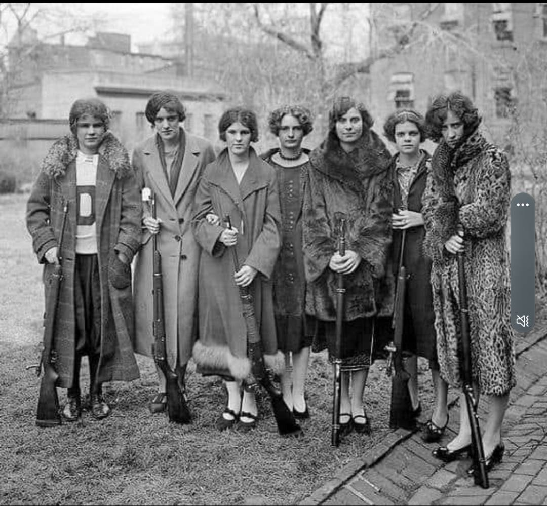 Seven women in 1920s-style coats and dresses stand outdoors in a yard, each holding a rifle and looking at the camera. Leafless trees and brick buildings are visible in the background.
