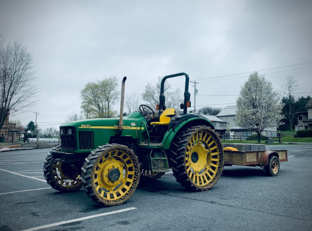 A green and yellow tractor with large, spoked wheels is parked in an empty lot, attached to a small wooden trailer. Trees and buildings are visible in the background under a cloudy sky.