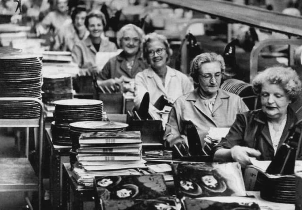 A group of women working in a factory assembly line, smiling while handling vinyl records and album covers. The workspace is filled with stacks of records and albums. The photo appears to be from the mid-20th century.