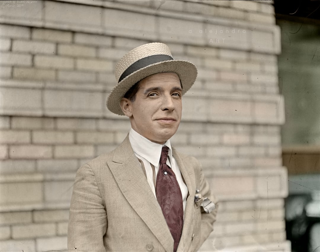 A man wearing a straw boater hat, light-colored suit, white shirt, and patterned tie stands in front of a brick wall, looking slightly to the side with a subtle smile.