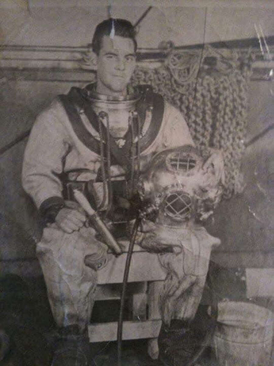 A man in a vintage deep-sea diving suit sits on a bench, holding a metal diving helmet. Heavy ropes hang in the background. The black-and-white photo gives an old-fashioned, historical feel.