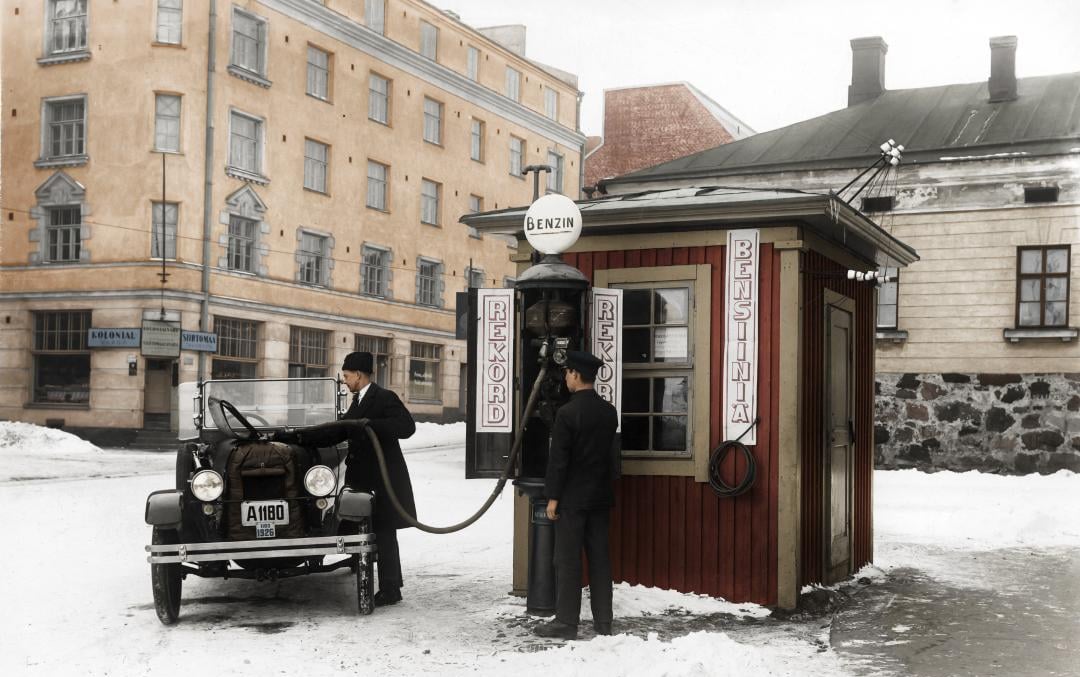 Two men stand by an old-fashioned car at a small, red gas station with a "BENZIN" sign, refueling the car. Snow covers the ground, and historic buildings are in the background.