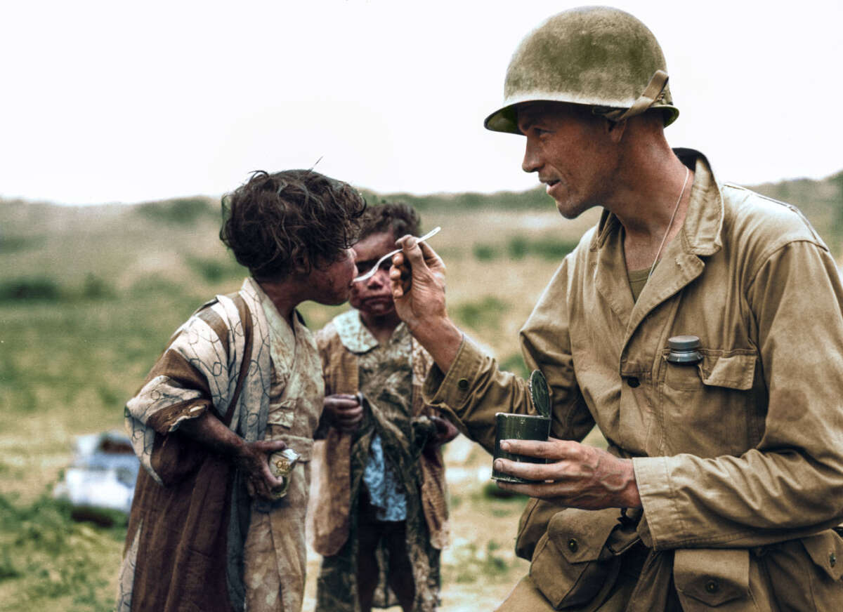 A soldier in uniform gently feeds a young child with a spoon, while another child stands nearby. The background is an open field, and the children appear dirty and wearing tattered clothes.