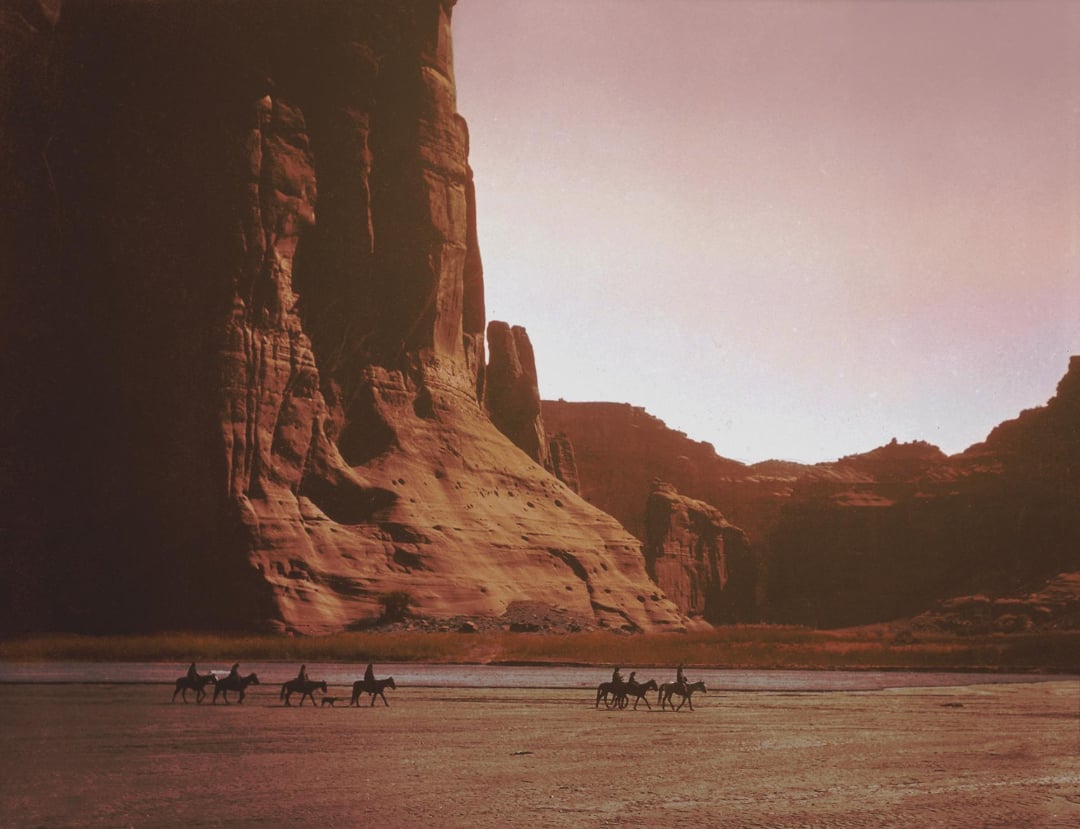 A group of riders on horseback travel across a wide, flat plain at the base of towering red sandstone cliffs under a hazy sky, with dramatic shadows cast across the landscape.