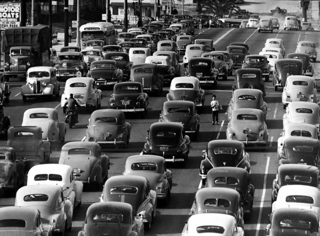 Black-and-white photo of heavy traffic with rows of vintage cars at a standstill. Two traffic officers direct vehicles at a busy urban intersection, and buildings are visible in the background.