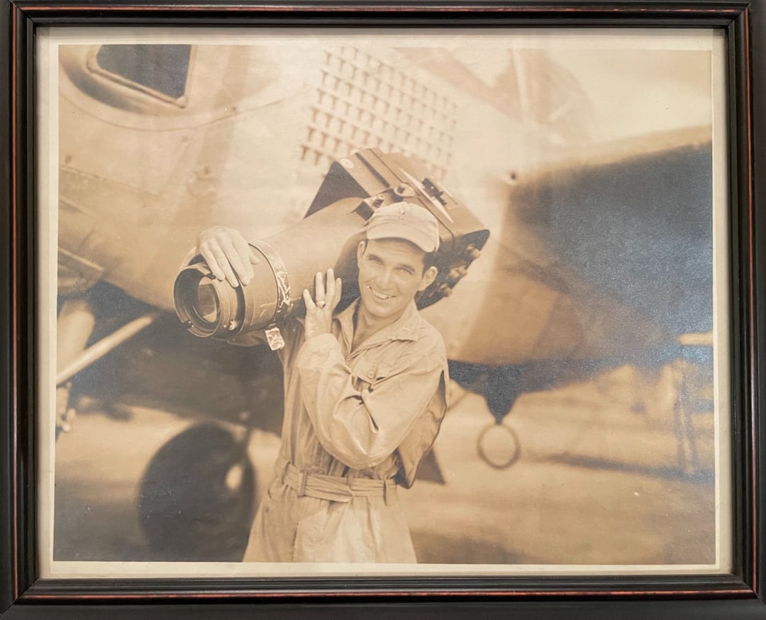 Sepia photo of a smiling man in a jumpsuit and cap, holding a large camera, standing beside an old airplane. The image is framed with a dark wooden border.