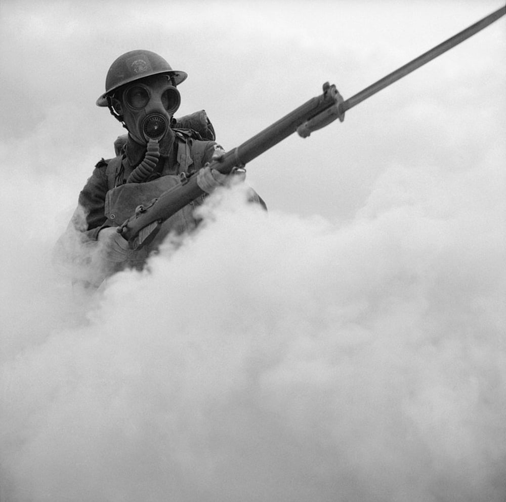 A soldier wearing a gas mask and helmet stands in thick smoke, holding a rifle with a bayonet attached.