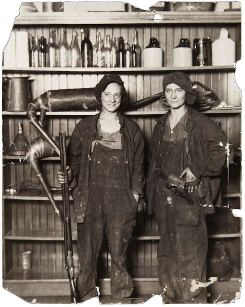 Two women in work overalls and hats stand in front of shelves filled with bottles and equipment, smiling at the camera; one holds a large tool, the other has tools at her waist. The photo is old and partially torn.