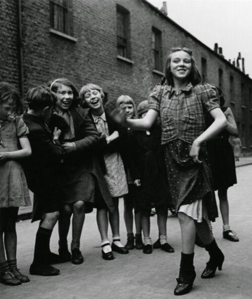 A group of smiling children, mostly girls in dresses, stand in a line watching and laughing as one girl dances joyfully in the street of a brick row house neighborhood.