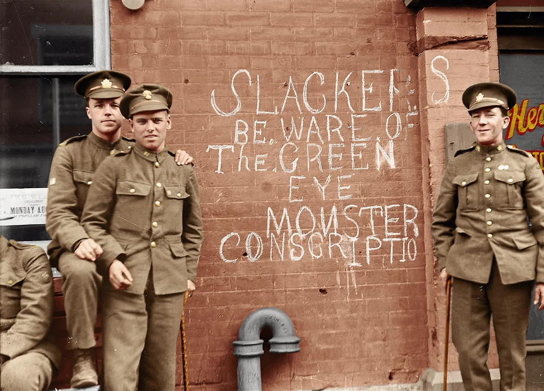 Four soldiers in uniform stand and sit in front of a brick wall with graffiti reading “SLACKERS BEWARE OF THE GREEN EYE MONSTER CONSCRIPTION.” One door and a window are visible behind them.