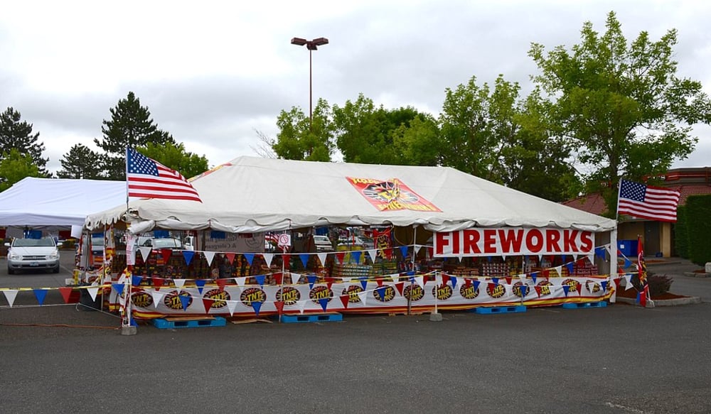 A large white tent decorated with American flags and colorful banners is set up in a parking lot, selling fireworks. The word "FIREWORKS" is prominently displayed on the tent. Trees and a cloudy sky are in the background.