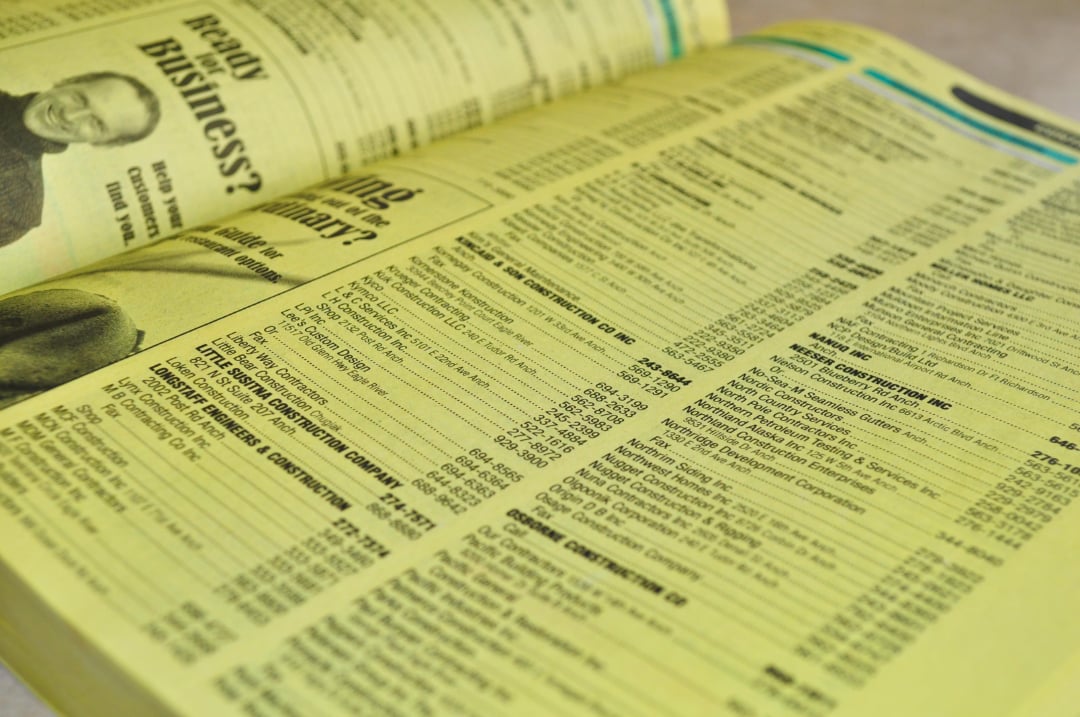 A close-up of a yellow phone book shows business listings and advertisements printed in black text on the pages. The directory is open, revealing several columns of contact information.