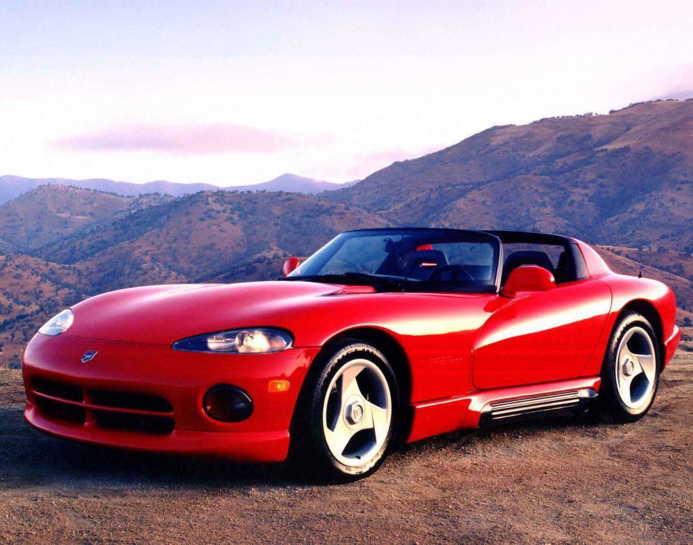 A red sports car is parked on a dirt road overlooking scenic, mountainous terrain under a partly cloudy sky.