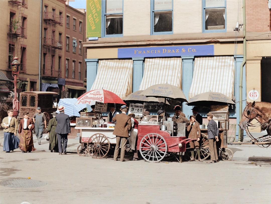 A bustling street scene in front of Francis Draz & Co., with people gathered around food carts. Horses and carriages are visible, and striped awnings cover storefront windows in the background.