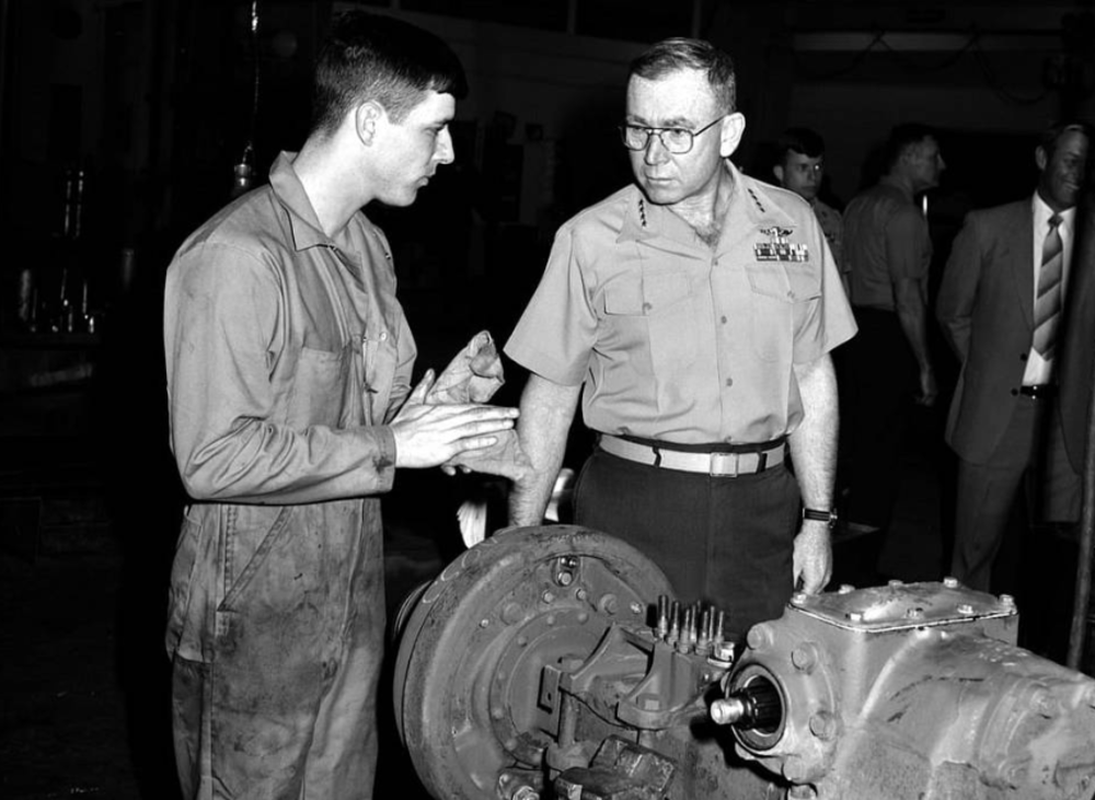 A young man in work overalls discusses machinery with a uniformed military officer in a workshop. Other men, some in military attire and others in suits, stand in the background observing the conversation.