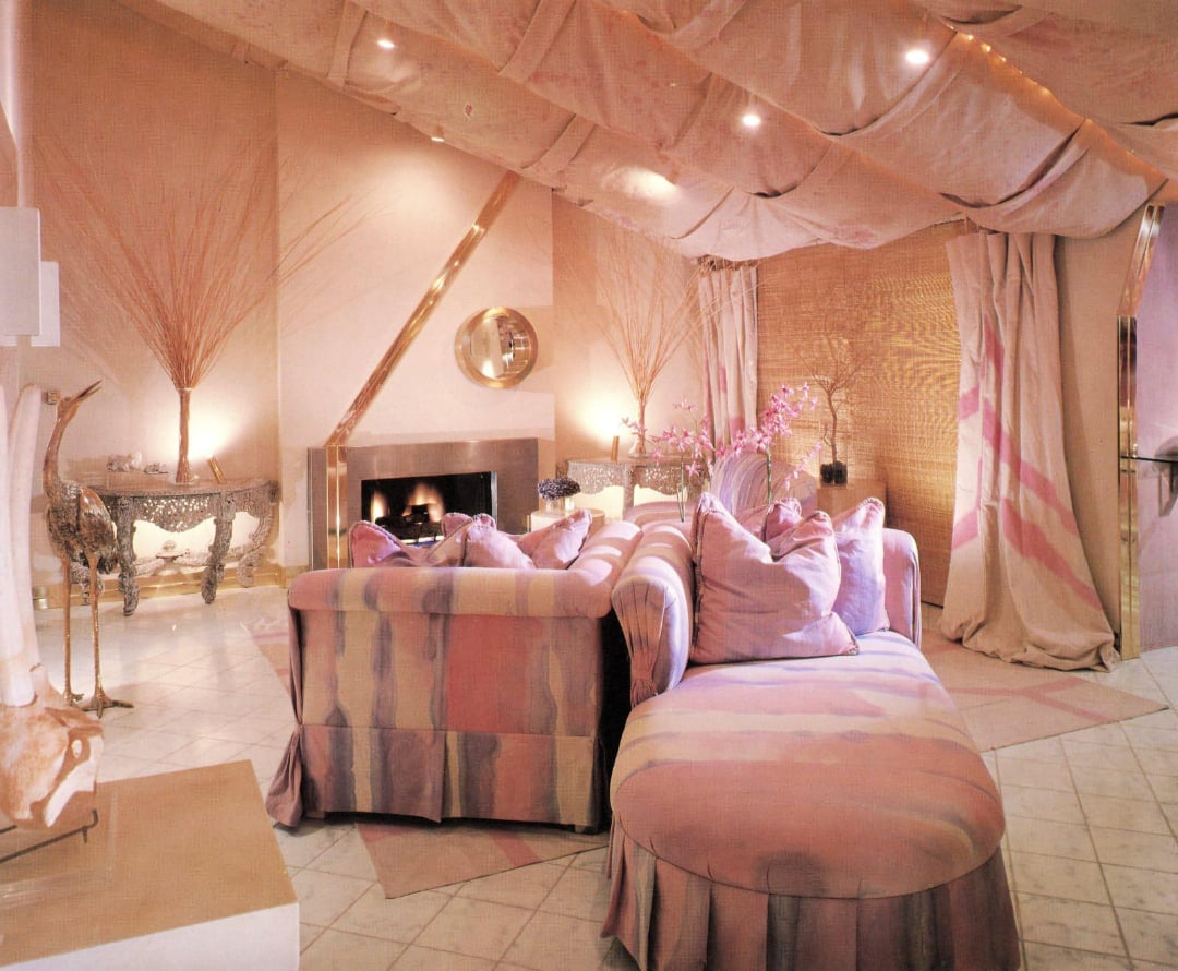 A pastel pink living room with draped ceiling, patterned sofa and chaise lounge, a lit fireplace, mirrored decor, and soft lighting creating a cozy, elegant atmosphere.