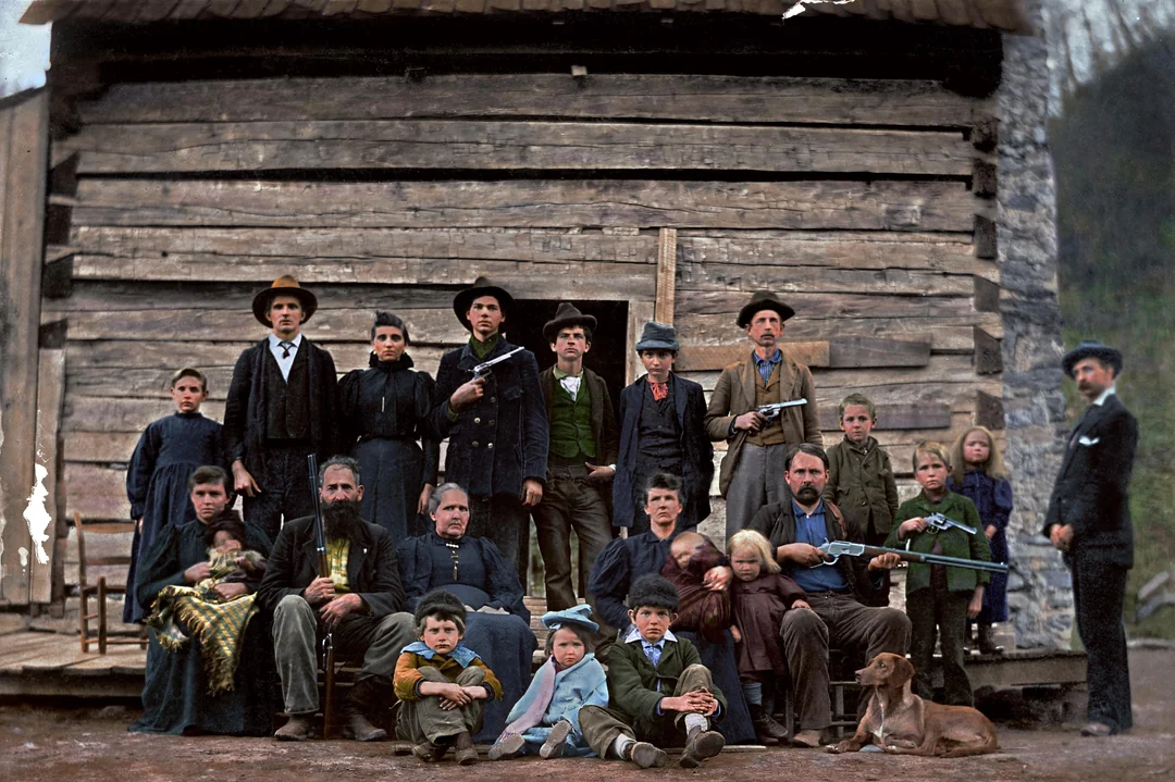 A large group of men, women, and children pose in front of a rustic wooden cabin; some men hold rifles, and a brown dog lies on the ground near the seated children.