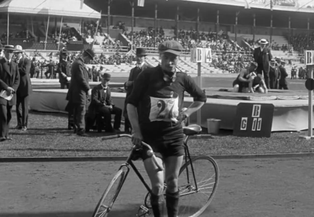 A vintage black-and-white photo of a male cyclist standing next to his bicycle on a track, wearing an old-fashioned sports outfit and cap, with a stadium and spectators visible in the background.