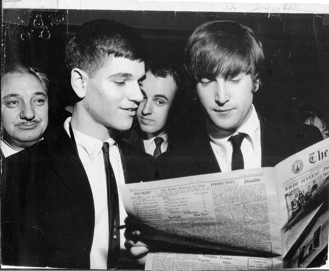 Three men in suits look at a newspaper together at a formal event. The man in the foreground on the right holds the newspaper, while the other two closely observe. All have serious or focused expressions.