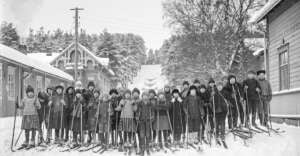 A large group of children dressed in winter clothes stand on a snowy street with skis and poles, flanked by wooden buildings and trees, posing for a photo in a historic, winter village setting.