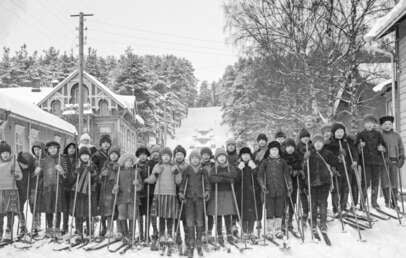 A large group of children dressed in winter clothes stand on a snowy street with skis and poles, flanked by wooden buildings and trees, posing for a photo in a historic, winter village setting.