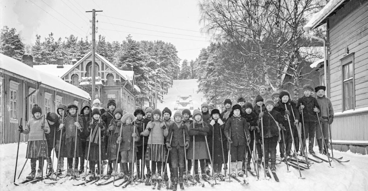 A large group of children dressed in winter clothes stand on a snowy street with skis and poles, flanked by wooden buildings and trees, posing for a photo in a historic, winter village setting.