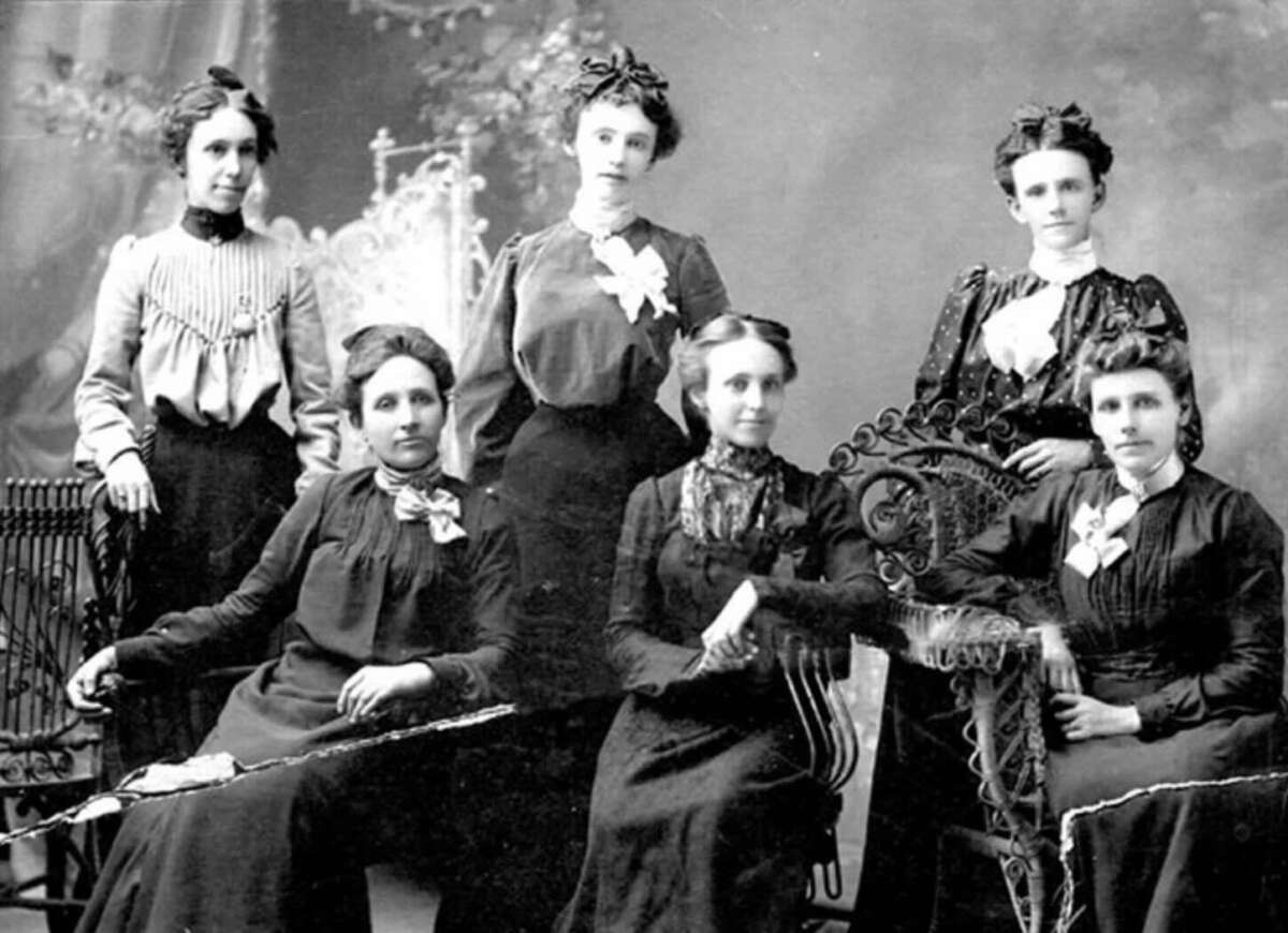 Six women in Victorian-era clothing pose for a formal black-and-white portrait, with three seated on ornate chairs and three standing behind them. The background features a decorative screen and painted scenery.