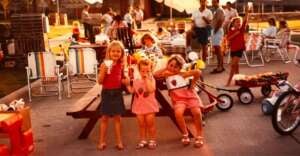 Three young girls, dressed in summer clothes, pose and smile in front of a picnic table at a lively outdoor gathering. Families, lawn chairs, and children's toys surround them on a sunny day.