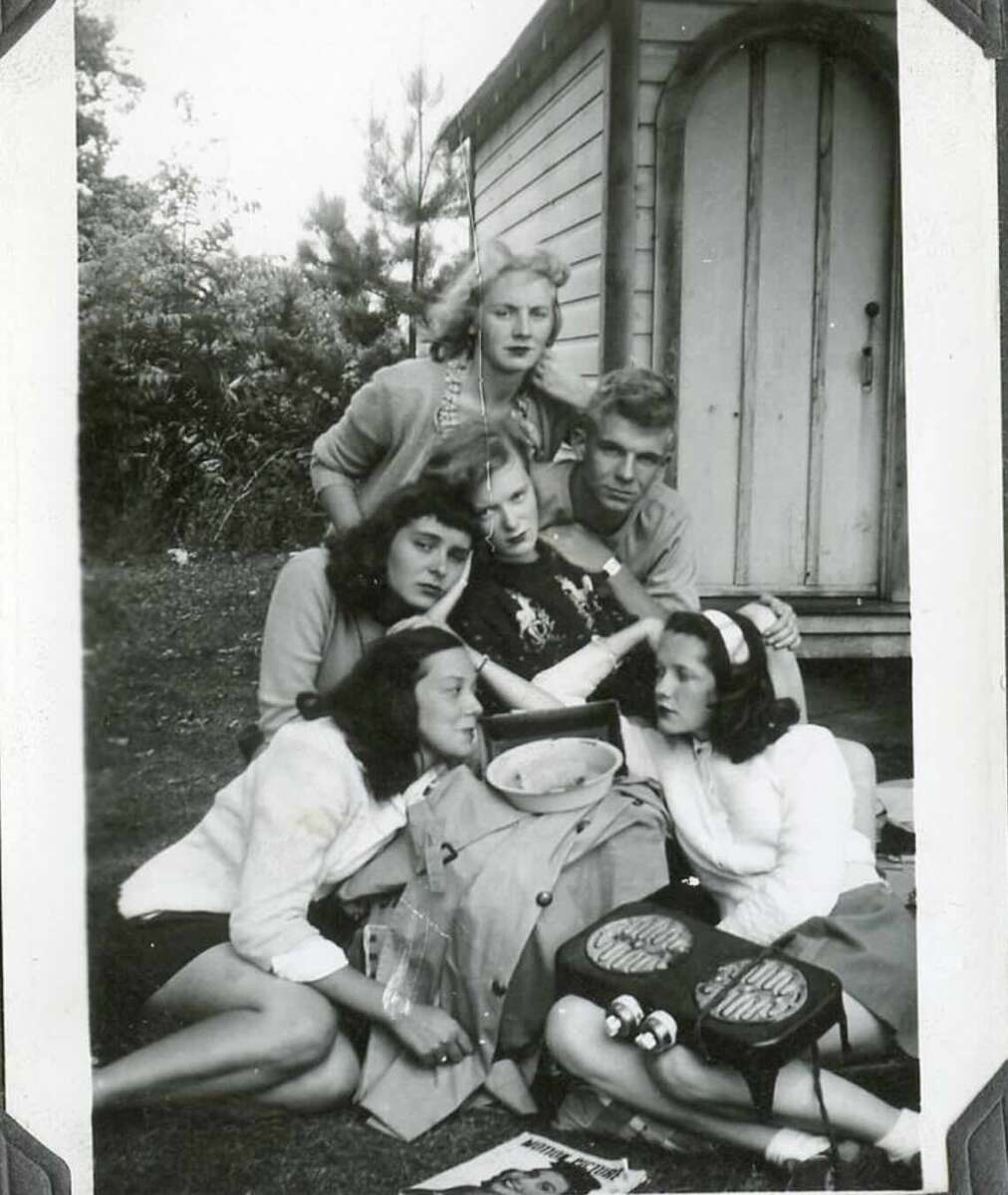 Six young people pose closely together outdoors in front of a small wooden shed. Some are sitting on the grass with bags and magazines, while others kneel behind them, smiling and looking at the camera.