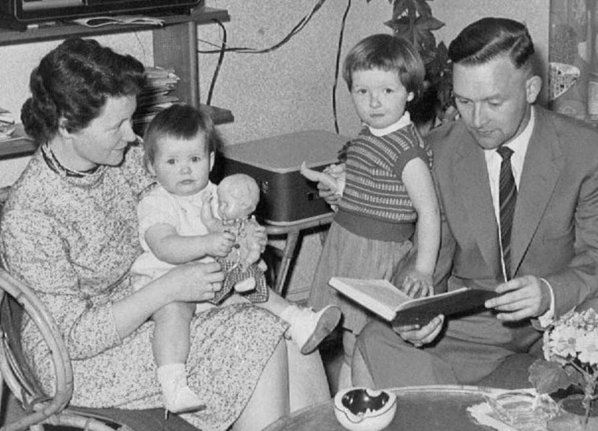 A black-and-white photo of a family: a woman holding a baby, a young child standing by her, and a man in a suit reading from a book. They are seated in a cozy, furnished room.