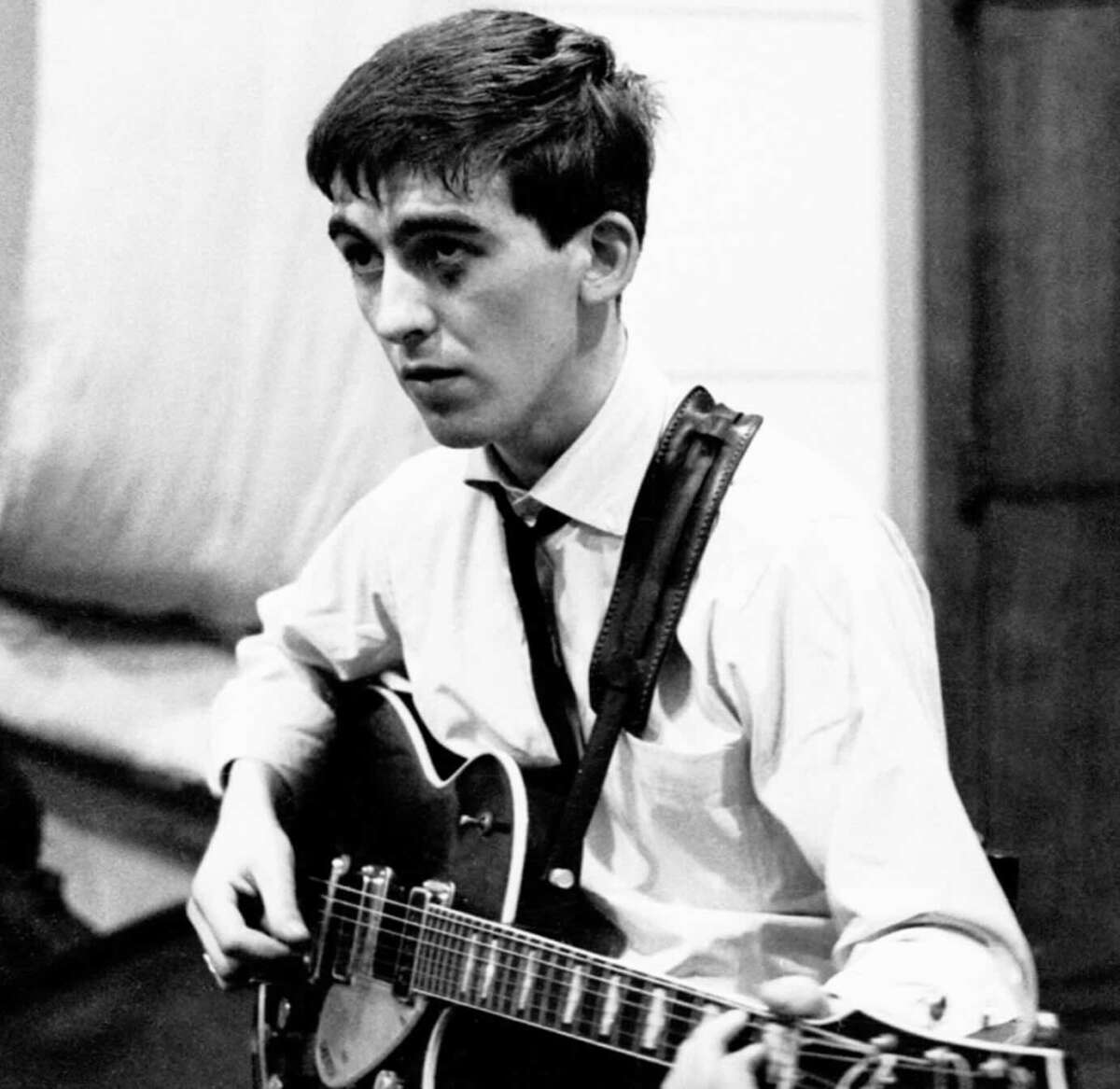 A young man with short dark hair plays an electric guitar. He is wearing a white shirt and a tie, sitting indoors, and appears focused, looking slightly to the side.