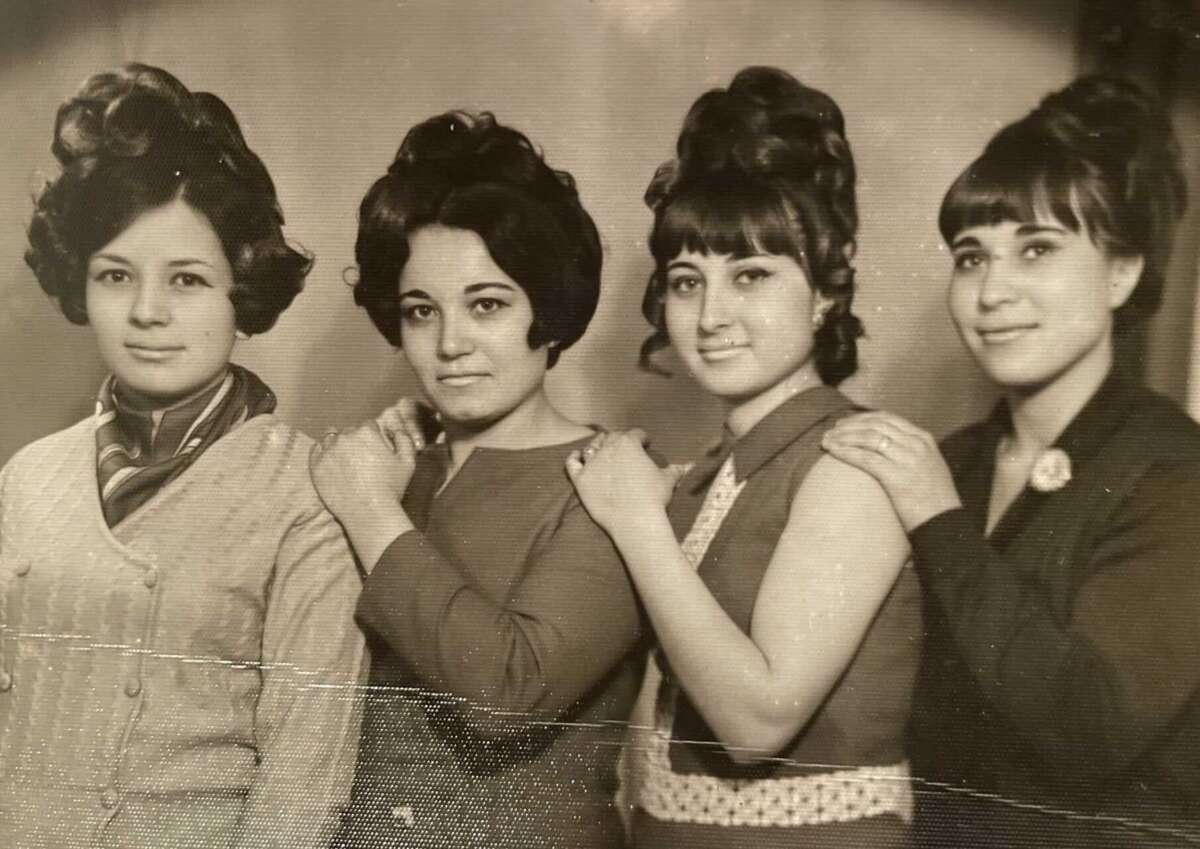 Four women with vintage hairstyles and clothing stand in a row, each resting a hand on the shoulder of the person in front. The photo is black and white, evoking a retro or 1960s-1970s era.