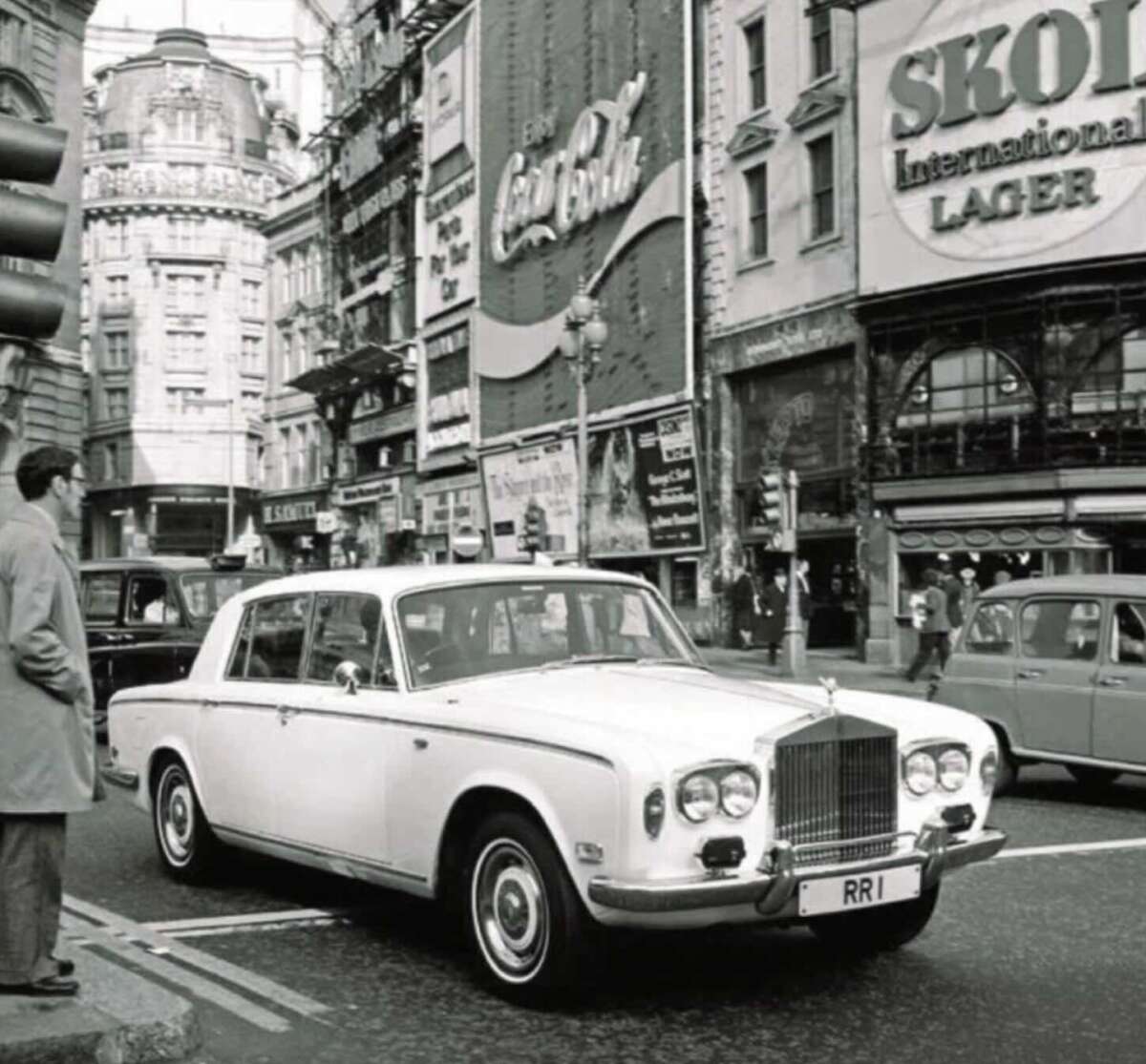 A classic white Rolls-Royce with the license plate "RR1" is parked on a busy city street, surrounded by vintage cars and large advertising billboards, including Coca-Cola and Skol Lager. The scene appears to be from the mid-20th century.