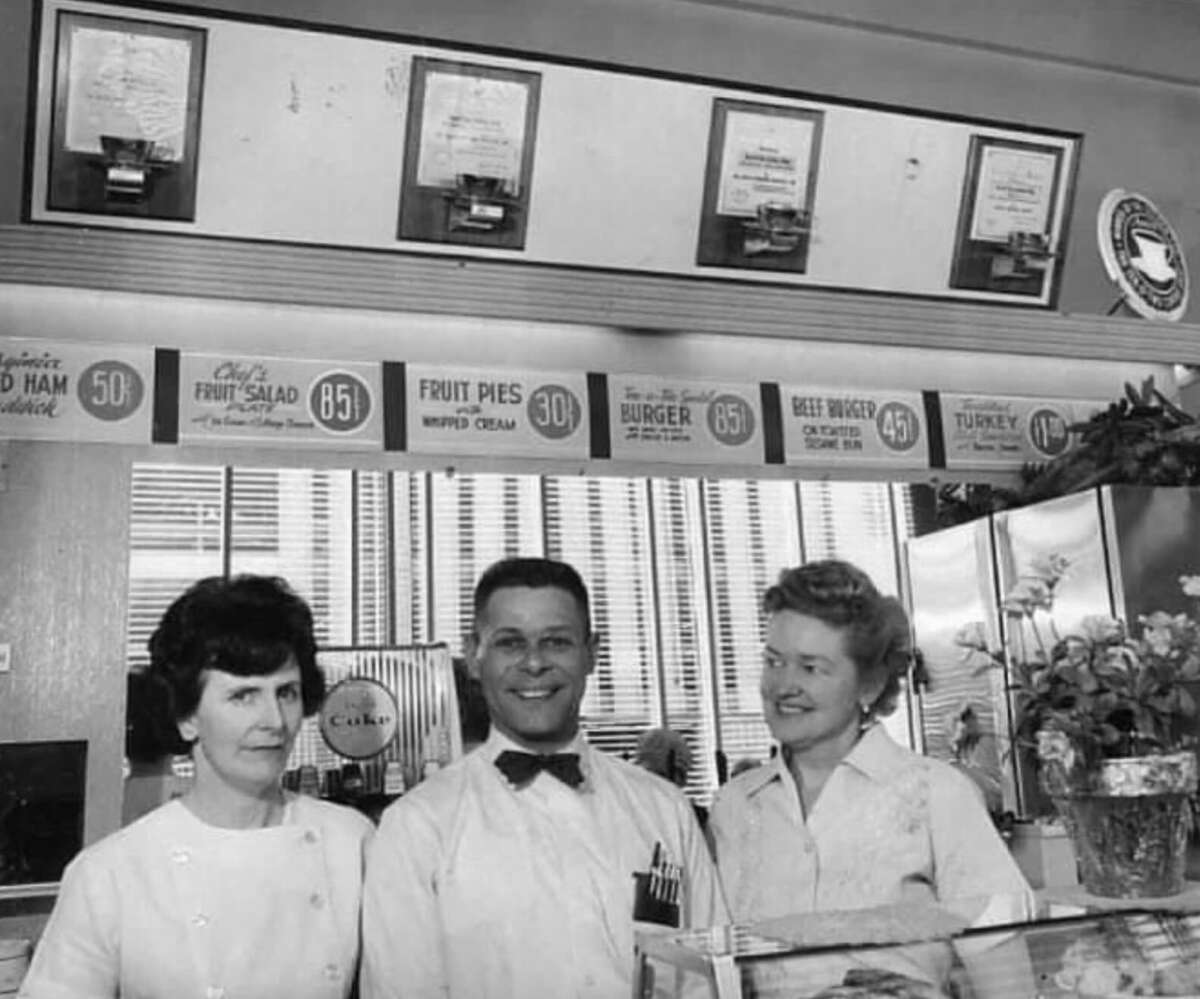 Three adults stand behind the counter of a vintage diner or cafe. Menu signs and framed certificates hang on the wall above them. The man in the center wears a bow tie and glasses; the women stand on either side, smiling.