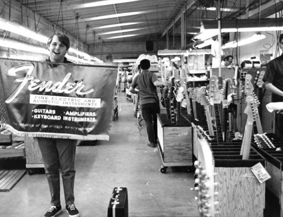 A smiling person holds a large Fender banner in a factory, with rows of electric guitar necks and bodies in stands. Workers assemble guitars in the background under fluorescent lights.