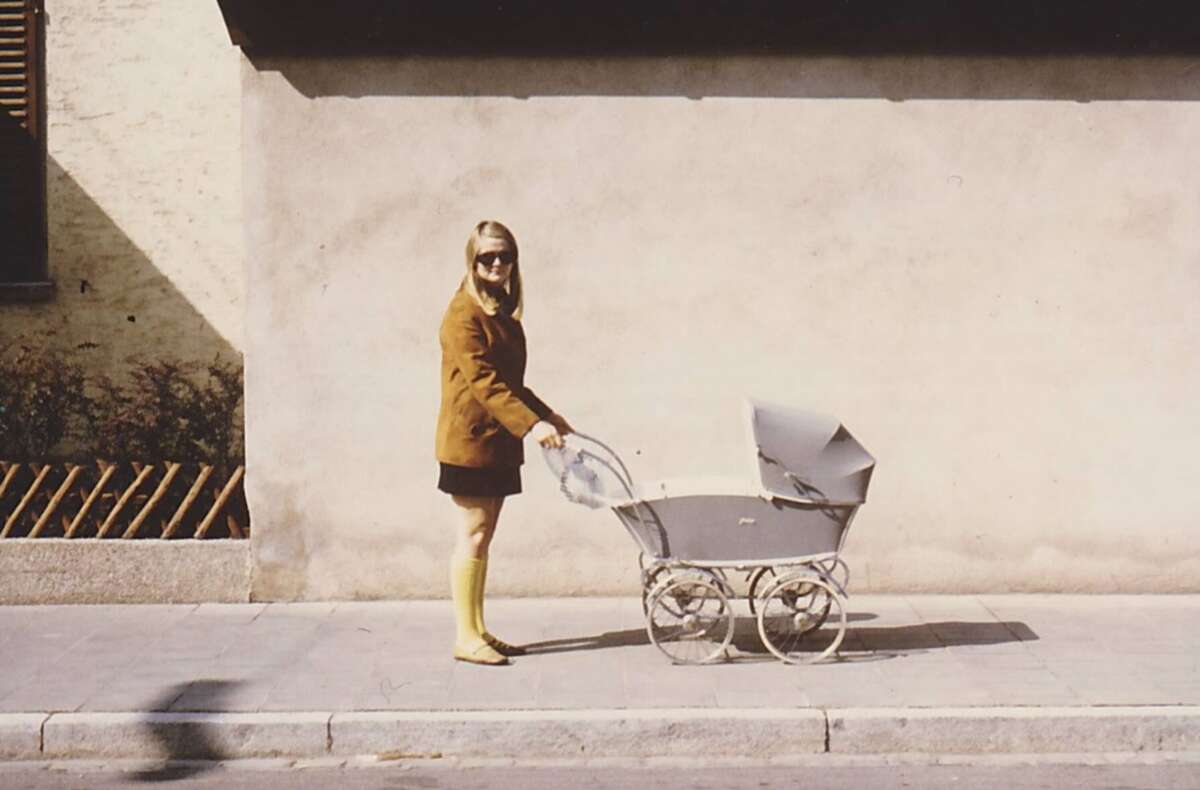 A woman wearing a brown jacket, black skirt, and yellow knee-high socks walks a vintage baby stroller along a sidewalk beside a beige wall.