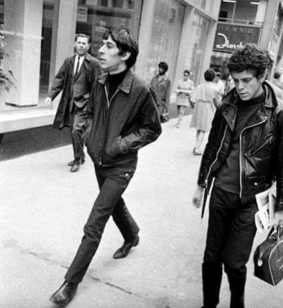 Two young men walk down a city street; one wears a zip-up jacket and slacks, the other a leather jacket and holds magazines. Pedestrians and shops are visible in the background, suggesting a busy urban setting.