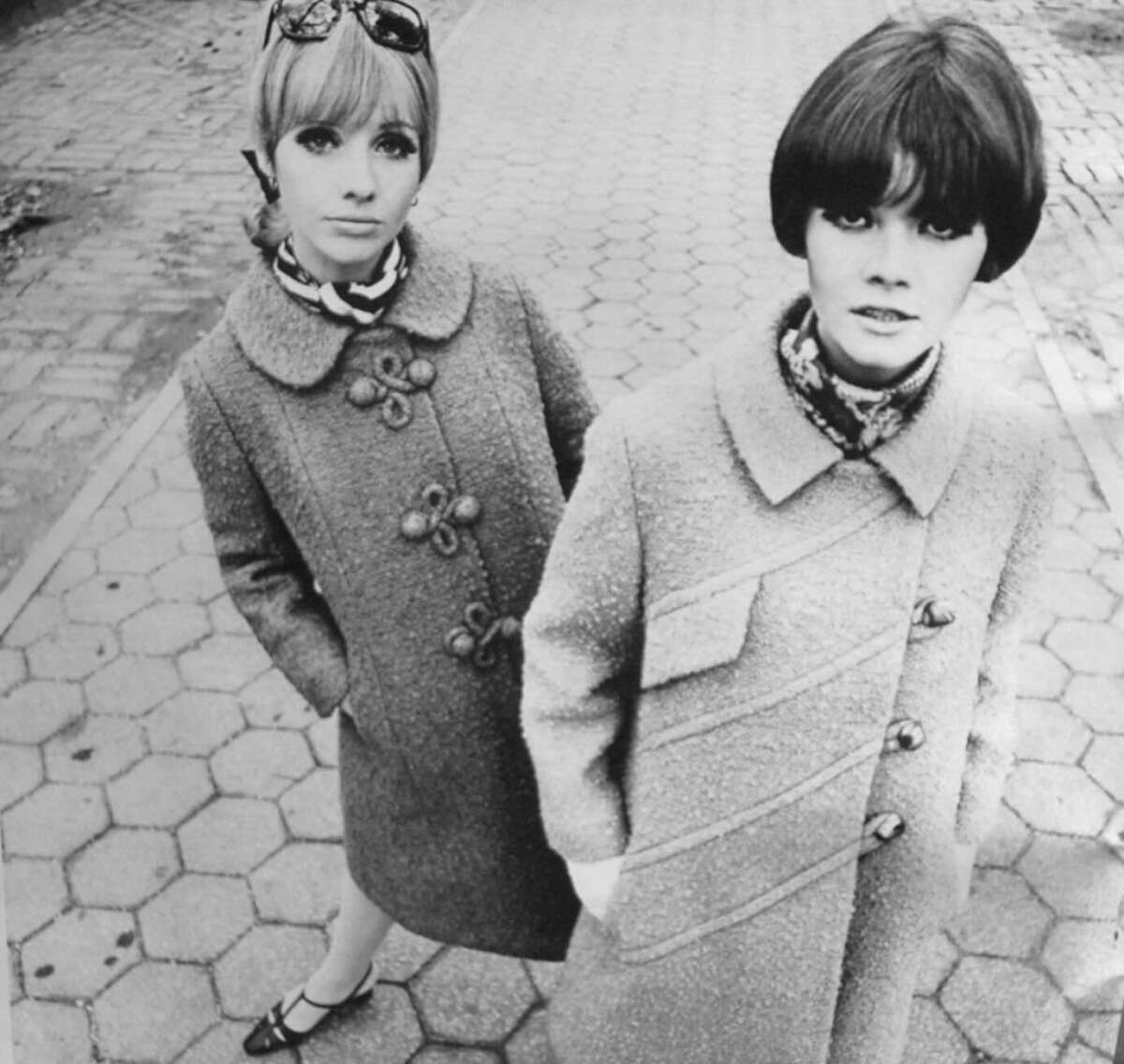 Two women wearing textured coats, patterned scarves, and short hairstyles stand on a hexagonal-tiled sidewalk. One woman wears sunglasses on her head and stands slightly behind the other. The image is in black and white.