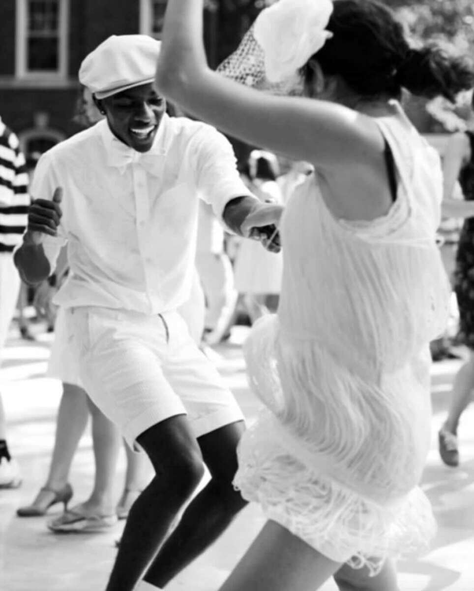 Two people dressed in vintage-style white outfits dance energetically together outdoors, appearing joyful. Other people in similar attire are seen in the background. The image is in black and white.