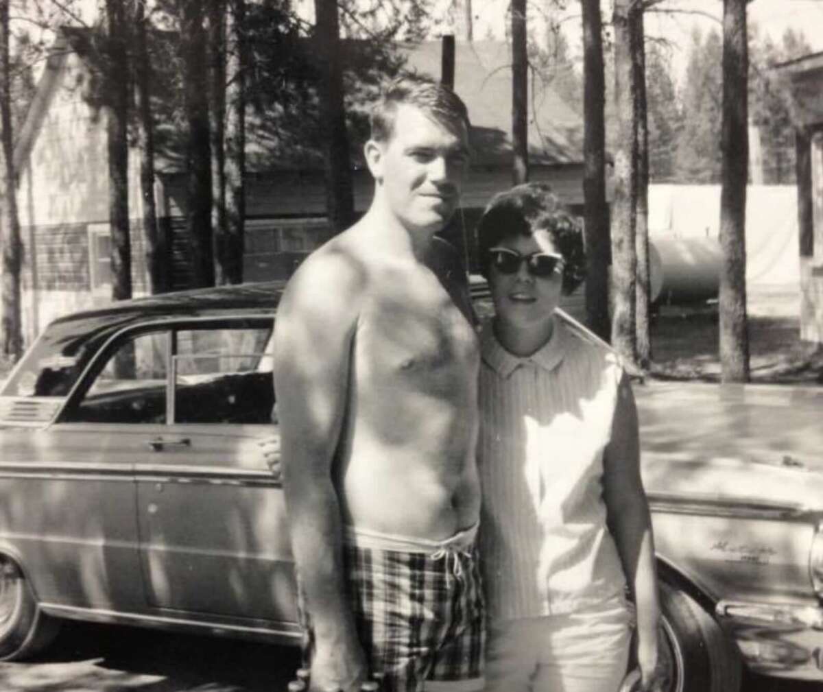 A man in swim trunks and a woman in sunglasses and a sleeveless blouse stand together in front of a vintage car, with trees and a house in the background. The photo is black and white.