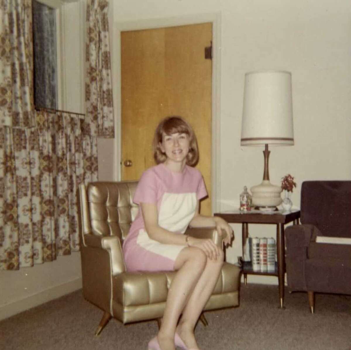 A woman with shoulder-length hair sits on a gold chair in a vintage living room, wearing a pink and white dress and pink shoes. Behind her are patterned curtains, a wooden door, a lamp, and a side table with magazines.