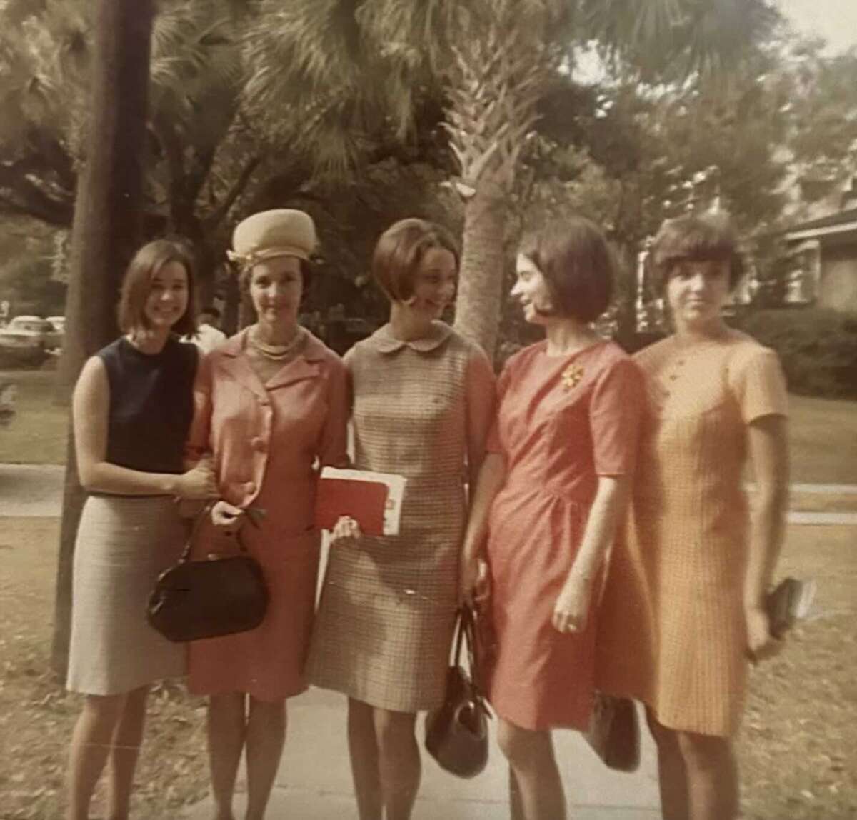 Five women stand together outdoors on a sidewalk, dressed in 1960s-style dresses in various colors. Palm trees and houses are visible in the background on a sunny day.