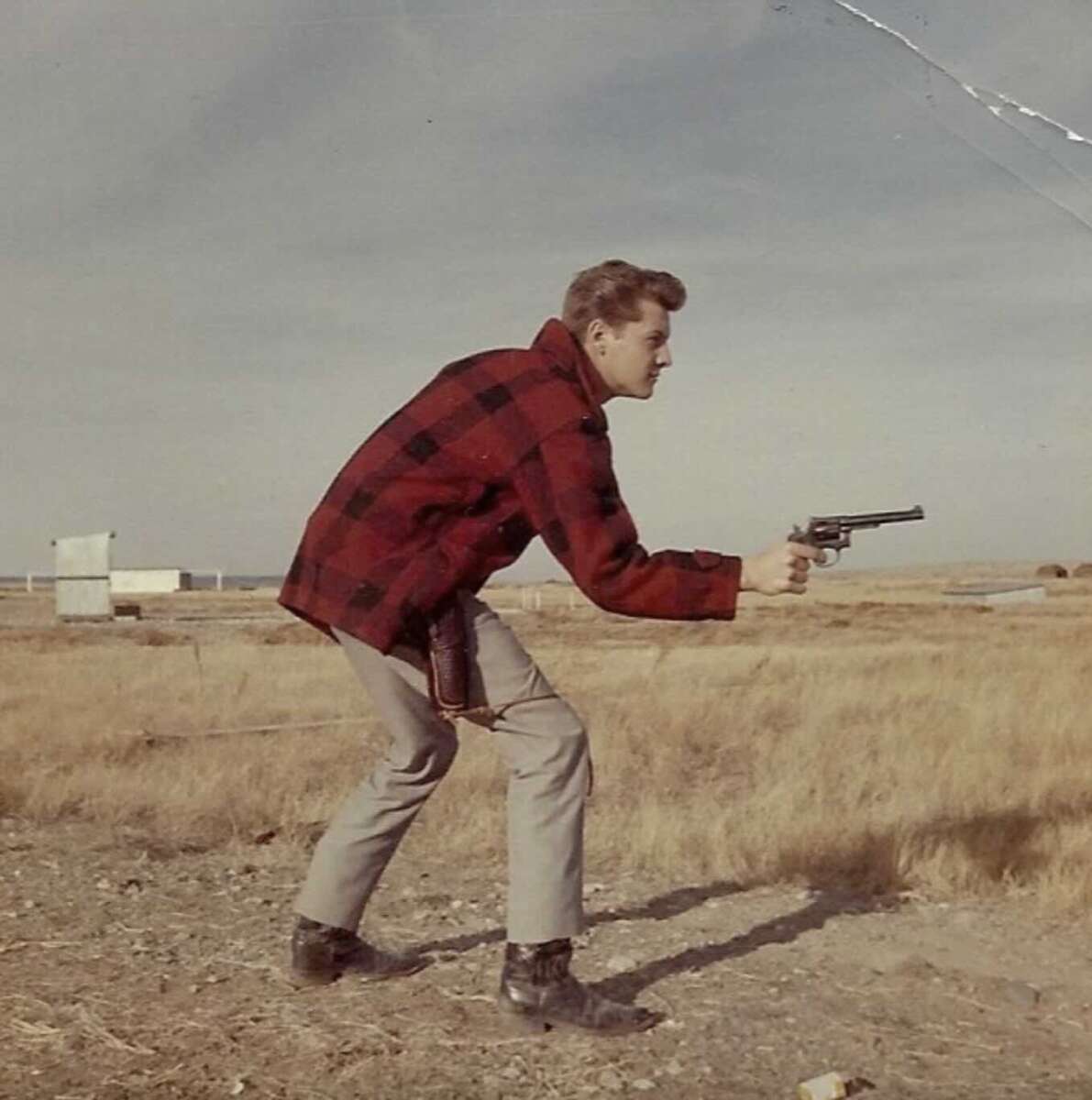 A young man in a red plaid jacket and gray pants crouches outdoors on dry grass, aiming a revolver with both hands under a cloudy sky, evoking a Western or cowboy scene.